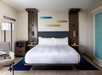 Modern hotel room with a large bed, two nightstands, a chair, a floor lamp, and colorful wall art above the headboard.
