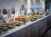 A buffet setup with various dishes displayed in chafing dishes and trays on a long countertop, including meats, salads, and desserts.