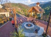 People sit and stand around a circular outdoor fire pit on a stone patio with mountains and trees in the background, near a small wooden building.
