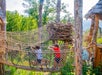 Children walk across a rope bridge surrounded by trees, with a wooden structure resembling a nest visible in the background.