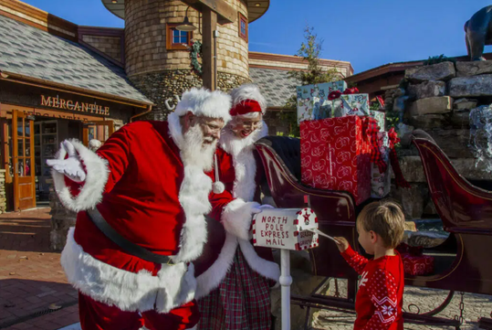 A boy in a red sweater places a letter in a "North Pole Express Mail" box while two people dressed as Santa and Mrs. Claus look on beside a sleigh with gifts.