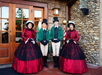 Four people in Victorian-style holiday costumes stand smiling outside a rustic building with wooden doors and a stone wall.