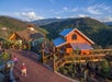 Two people walk on a brick pathway past wooden buildings overlooking forested mountains under a clear blue sky.
