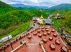 Outdoor patio with round tables and chairs overlooks a lush, mountainous landscape with buildings and winding roads in the background under a cloudy sky.