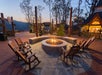 Outdoor fire pit with a ring of wooden chairs on a stone patio, surrounded by shops and trees at dusk.
