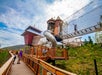 An elevated wooden play structure with towers, a slide, and a rope bridge is surrounded by greenery; people walk on the adjacent wooden path.