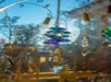 A multicolored crystal prism ornament hangs in sunlight, reflecting blue and purple hues, with trees and a wooden structure blurred in the background.