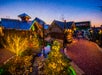 Village scene at dusk with Christmas lights on trees and buildings, a small stream, and people walking along a pathway.