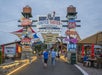 People walk through a lively outdoor food court area with colorful signs, including "United Tastes of America" and "Route 66," under a cloudy sky.