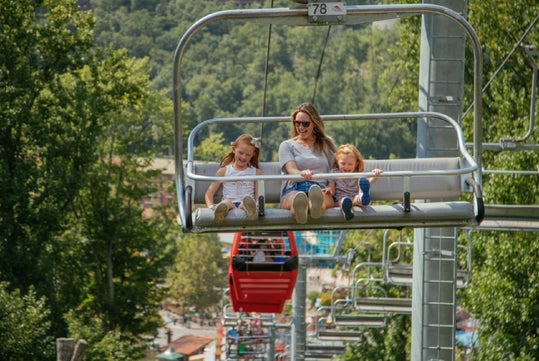 A woman and two children ride a chairlift above a green forested area and a red gondola, all seated and smiling.