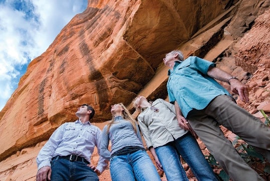 Four people stand on rocky ground, looking up at a large red rock formation under a partly cloudy sky.