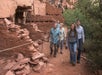 Five people walk along a dirt path past ancient red stone ruins and vegetation in a desert setting.