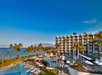 Resort with modern buildings, pools, and palm trees near the ocean, under a clear blue sky.