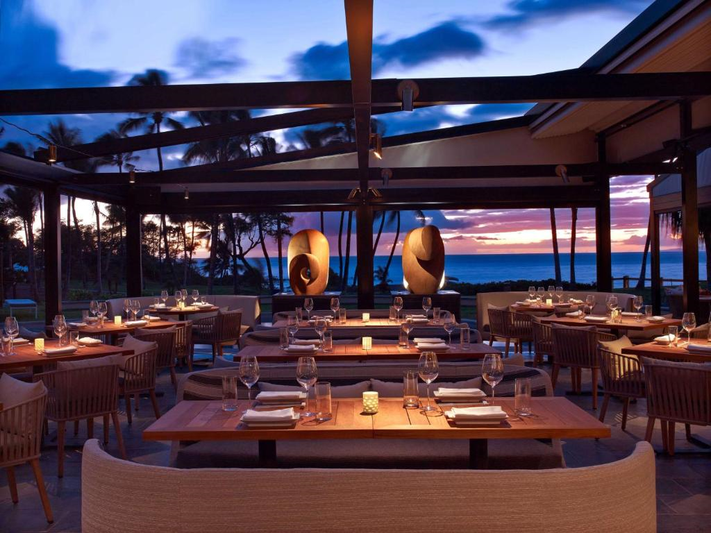 Outdoor restaurant with wooden tables and cushioned chairs, set for dining at sunset. Sculptures and palm trees are visible, with the ocean in the background.