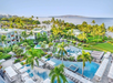 Aerial view of a tropical resort with multiple pools, palm trees, green lawns, and the ocean in the background under a clear sky.