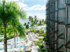 View of a hotel pool area with lounge chairs, palm trees, and the ocean in the background on a sunny day.