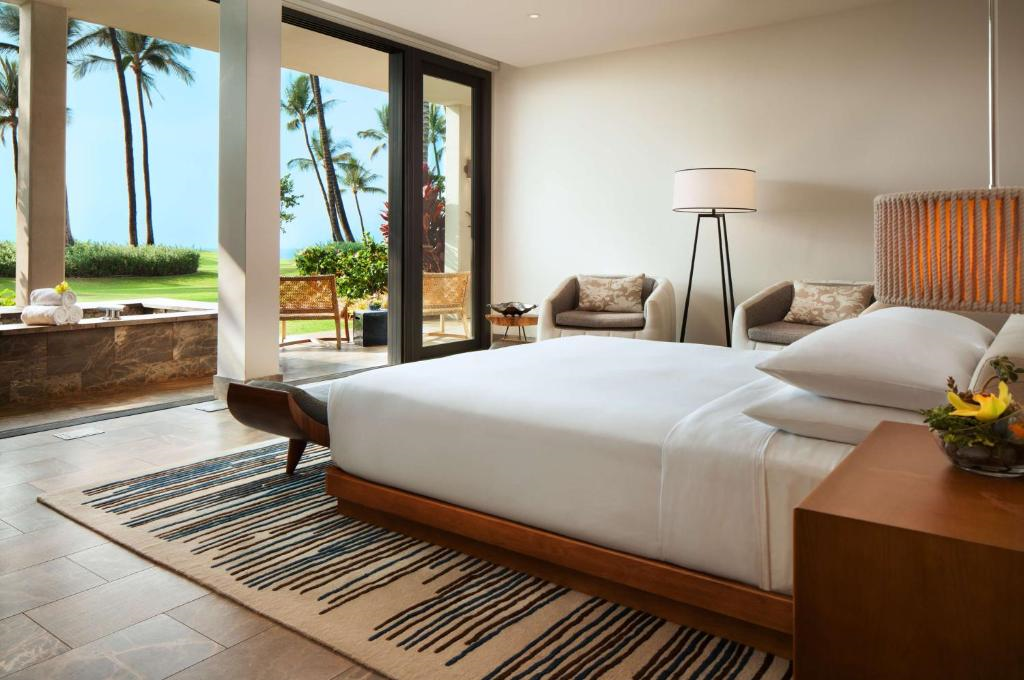 Hotel room with a large bed, wooden headboard, and striped rug. Floor-to-ceiling windows open to a patio with chairs and view of palm trees and grass outside.