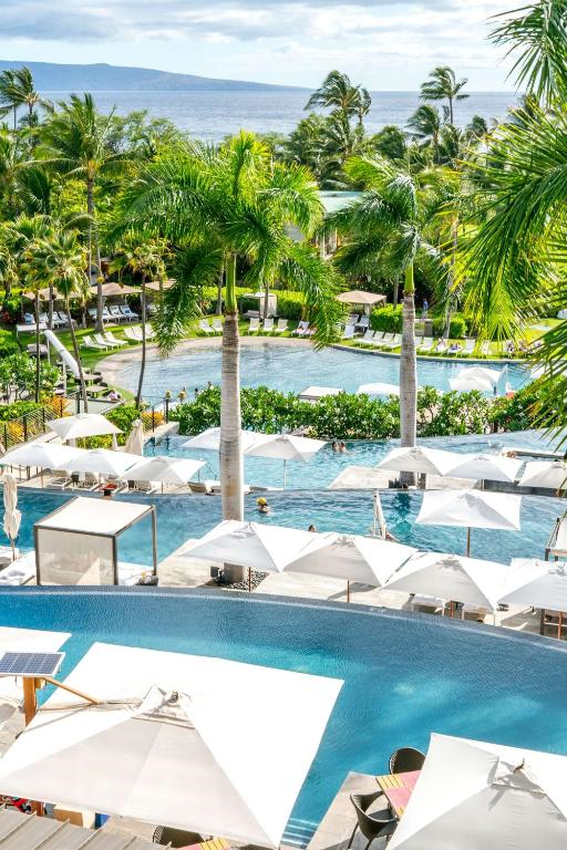 Aerial view of a tropical resort featuring multiple infinity pools, palm trees, sun loungers, and white umbrellas with the ocean visible in the background.