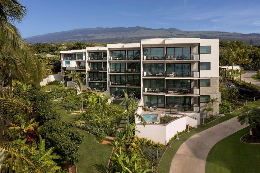 A modern multi-story apartment building with large windows, surrounded by lush tropical greenery, under a clear blue sky.