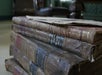 A close-up of a stack of old, worn books with faded leather covers and visible damage, resting on a surface with a piece of paper on top.