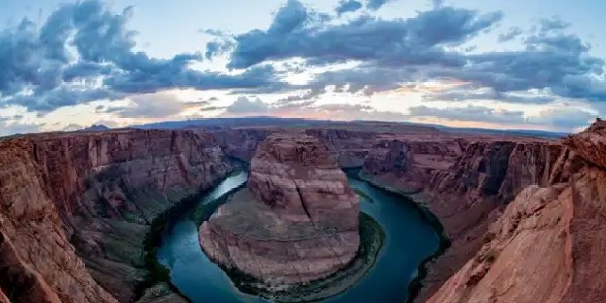 A wide view of Horseshoe Bend shows the Colorado River curving sharply around a rock formation, surrounded by steep red canyon walls under a partly cloudy sky.