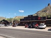 A rustic wooden building with a Coca-Cola sign, motorcycles, and cars parked in front, set against a desert landscape with hills and a clear blue sky.