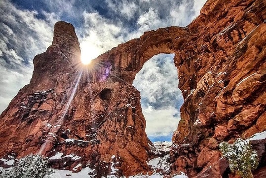 A natural rock arch rises against a partly cloudy sky, with the sun shining through the opening and patches of snow on the ground and rock surfaces.