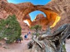 A group of people walk among rocky terrain and twisted trees beneath large natural stone arches at Arches National Park, Utah, under a clear blue sky.