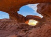 A natural rock arch frames the sunset and sky, with people walking along the rocky path beneath the arch in a desert landscape.