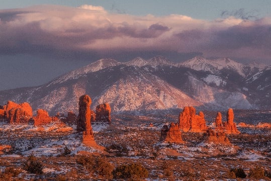 Red rock formations and snow-dusted desert landscape with mountains in the background under a cloudy sky at sunset.