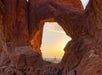 People stand at the base of a large natural rock arch at sunset, with sunlight illuminating the rock formations and the sky visible through the opening.