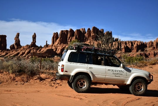 A white NAVTEC SUV driving on a red dirt road with the beautiful "The Needles" of the Canyonland as the background.
