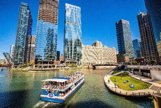 A tour boat travels on a river beside a cityscape with tall glass buildings. Sunny day with a clear blue sky.