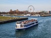 Tourist boat on a river with a crowded deck, cityscape and Ferris wheel in the background.
