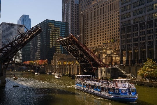 A tour boat passes under a raised drawbridge on a sunny day, surrounded by tall buildings and autumn trees.