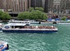 Tour boat on a river with passengers, passing by urban buildings and greenery.