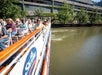 People are seated on a river tour boat passing by a building with green ivy and large windows.