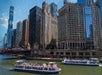 Boats with passengers navigate a city river, with tall buildings lining the waterfront under a clear blue sky.