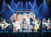 Theater performance of a nativity scene with actors in costumes, dancers in flowing dresses, and a background featuring stage lighting and props.