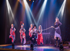 Performers on stage in colorful costumes; a man sits in the center while women in red dresses dance and one holds a lasso. Bright spotlights illuminate the scene.