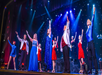 Performers in colorful costumes stand on stage, arms raised in a wave, with a starry backdrop and spotlighting.