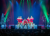 A person plays guitar on stage with four dancers dressed in festive outfits, surrounded by Christmas decorations and colorful lights.