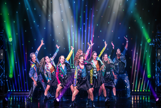 A group of nine performers in colorful jackets dance energetically on stage under bright lights with a starry backdrop.