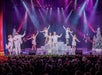 A group of performers in sparkling white costumes sing and dance on a brightly lit stage with Christmas trees, while an audience watches.