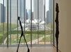 Two thin, abstract human sculptures stand inside a gallery with large windows overlooking an urban park and city skyscrapers.