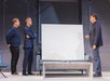 Three men stand and face each other in a modern room, with a blank white canvas propped on a couch between them and a marble coffee table in the foreground.