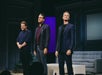 Three men stand on a stage in front of a sofa and tables, facing the audience, appearing to be at the end of a performance.