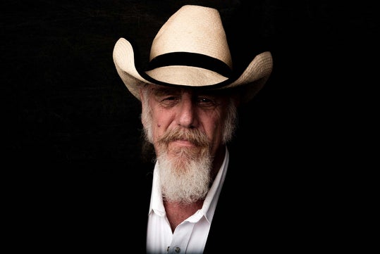A portrait of an older man with a long white beard wearing a cowboy hat, black suit jacket, and white shirt with a black background.