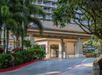 Entrance of a hotel with a covered driveway, surrounded by lush greenery and palm trees, leading to a multi-story building.