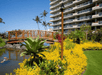 Lush garden with a wooden bridge over a pond, surrounded by vibrant yellow flowers and greenery, near a multi-story building under a clear blue sky.